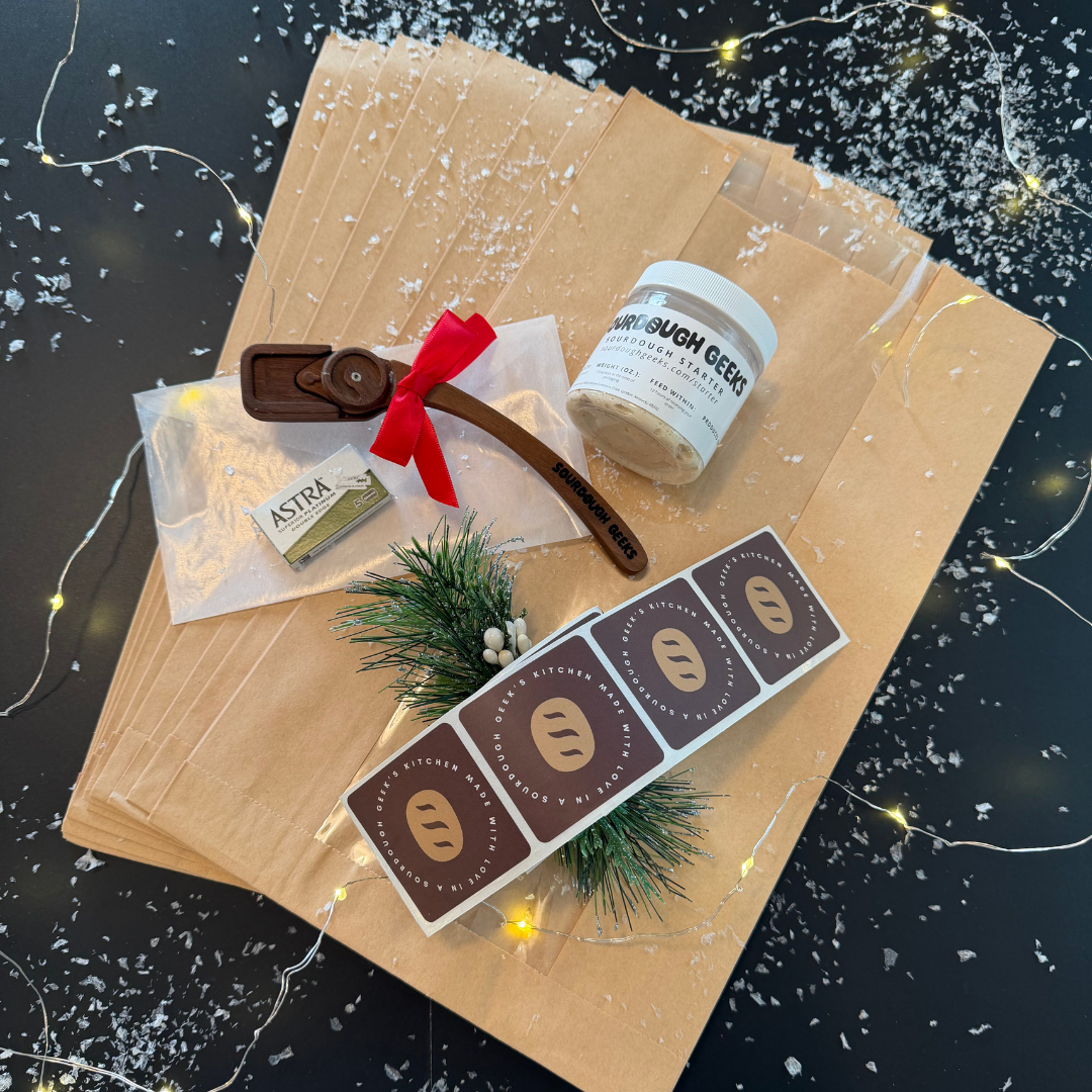 A bakers lame with razors, a jar of starter and a strip of stickers sit atop a dozen kraft bread bags, surrounded by snow and lights.